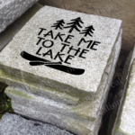 Engraved granite cottage accent stone feature with "Take Me To The Lake" and pine trees/canoe on stack of other granite stones.