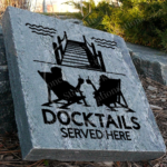 Engraved granite cottage/lake accent stone feature with "Docktails Served Here" and two people relaxing in chairs by dock having a drink near walkway.