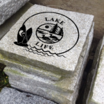 Engraved granite cottage/lake accent stone feature with "Lake Life" and pine trees, loons, paddleboards placed outside.